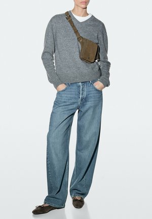 Persoon die een grijze trui, wit ondershirt, losse blauwe spijkerbroek, bruine schoudertas en donkerbruine ballerina's draagt, staand met handen in de zakken.