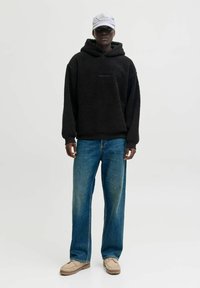 Schwarzer Fleece-Hoodie mit Logo auf der Vorderseite, kombiniert mit hellblauen weit geschnittenen Jeans und beigen Schuhen, vor einem neutralen Hintergrund.