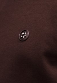 Στρογγυλή σκουρόχρωμη καφέ επιφάνεια με ανάγλυφο μεταλλικό λογότυπο "SB" ραμμένο σε σκουρόχρωμο καφέ ύφασμα.