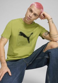 Camiseta de algodón verde con el logo negro de Puma sobre un patrón de cuadrícula; combinada con unos jeans oscuros desgastados. Mangas cortas y corte relajado.