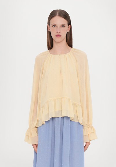 Light yellow sheer blouse with a rounded neckline, ruffled hem, and long sleeves; features subtle sparkle details. Paired with a blue skirt.