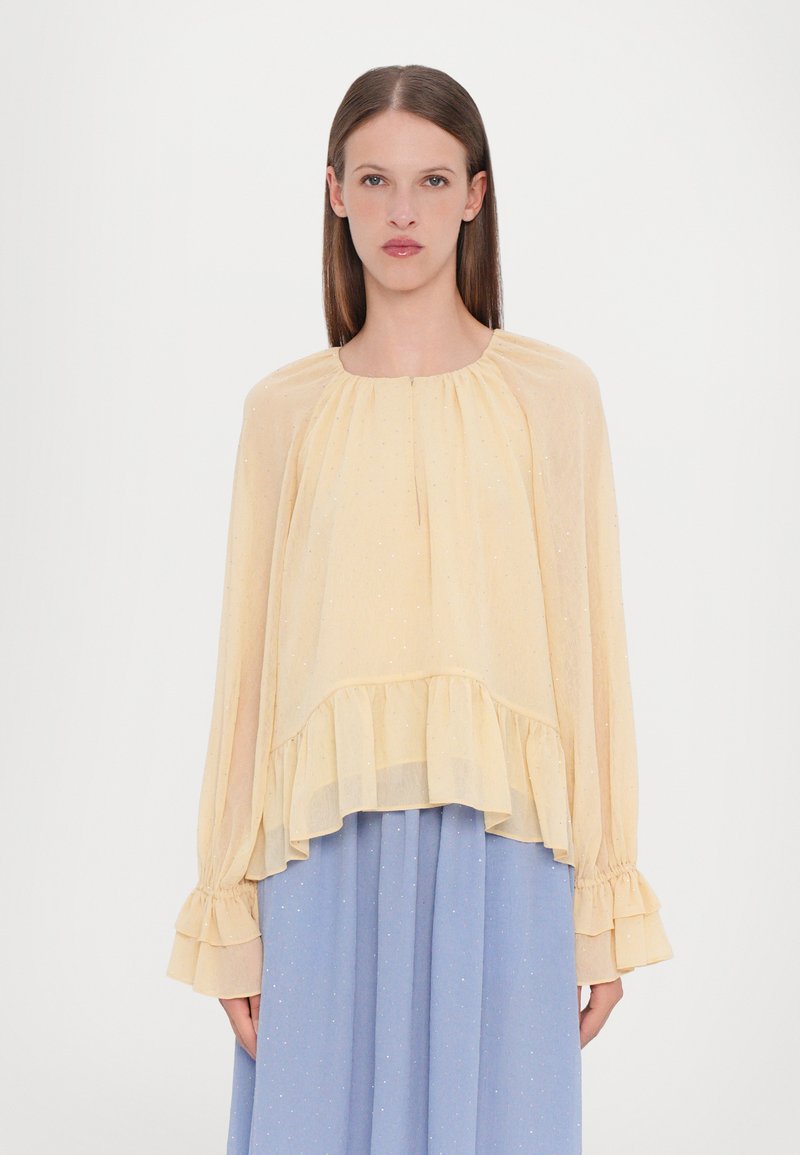Lichtgele doorschijnende blouse met een ronde halslijn, gerimpelde zoom en lange mouwen; bevat subtiele sprankelende details. Gepaard met een blauwe rok.