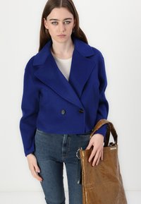 Blå dobbeltradet cropped jakke med store slag, parret med en hvid top og mørke jeans, mens der bæres en brun blank tote bag.