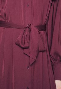 Burgundské šaty mají knoflíkový přední díl, s sklady a páskem s mašlí, vyrobené z hladkého, lesklého materiálu.