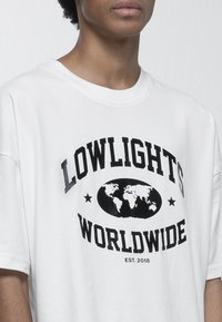Bílý bavlněný tričko s černým potiskem "LOWLIGHTS WORLDWIDE," grafikou zeměkoule a textem "EST. 2018." Krátké rukávy, kulatý výstřih.