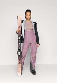 Lyžiarsky outfit pozostávajúci zo svetlomodrej bundy, vzorovaného purpurového topu a purpurových nohavíc s vysokým pásom a sivými akcentmi. Snowboard držený vertikálne.