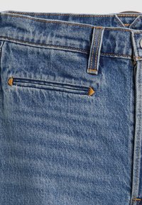 Gros plan sur un jean en denim bleu montrant une poche latérale avec une fente horizontale et un renforcement de couture jaune aux extrémités, une passants de ceinture et des détails de couture.