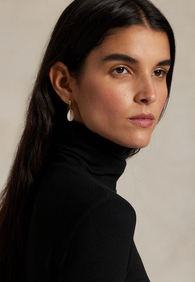 Pruunide pikkade juustega naine, kes kannab musta kõrge kaelusega kampsunit ja ühte valget kestakujulist kõrvarõngast, vaadates kergelt paremale tavalise tausta ees.