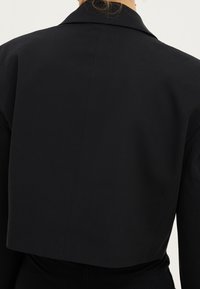 Bakifrån av en person som bär en figurnära svart kort jacka med bred krage och långa ärmar.