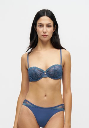 Frau mit langen dunklen Haaren, die einen blauen Spitzen-BH und passende blaue Unterhose trägt, steht vor einem einfachen hellen Hintergrund.