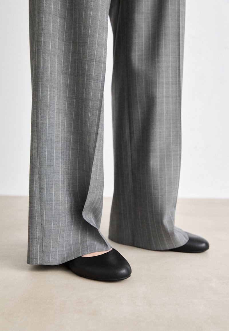 Jambes vêtues d'un pantalon large gris à fines rayures, associé à des chaussures plates noires à bout rond sur un sol clair.