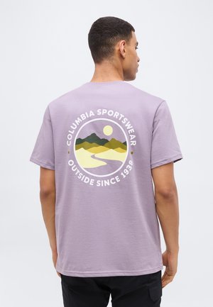 Mann trägt ein hellviolettes T-Shirt mit einer kreisförmigen Berg- und Sonnen-Grafik sowie dem Text "Columbia Sportswear Outside Since 1938" auf der Rückseite.
