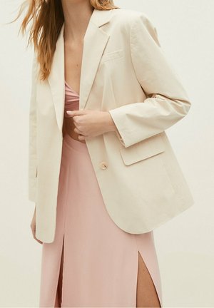 Mujer con blazer color crema sobre un bralette rosa claro y falda con abertura alta, de pie frente a un fondo liso y claro.