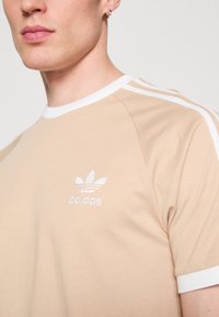Bežinė marškinė suapvaliu baltu apykaklė ir baltais šonų juostelėmis, su mažuoju baltu Adidas logotipu ant krūtinės. Minkšta medvilninė medžiaga.