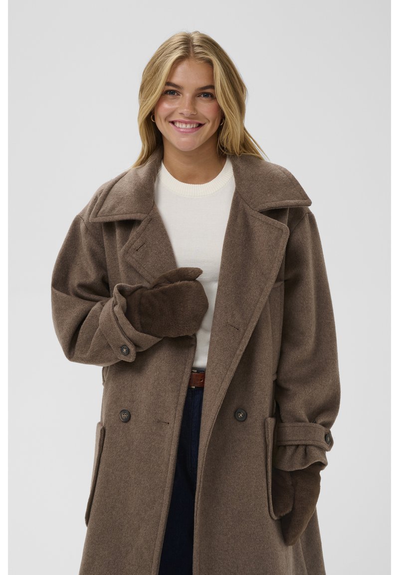 Šypsosi moteris su ilgais blondinų plaukais, vilkinti rudą vilnonį paltą, derančias pirštines, baltą megztinį ir tamsias kelnes, stovinti prieš paprastą foną.