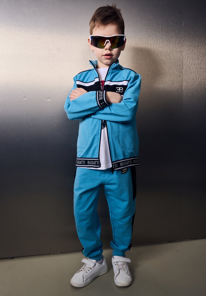 Ragazzo giovane in tuta blu Bugatti e sneakers bianche in piedi con le braccia incrociate, indossa occhiali da sole riflettenti con montatura bianca davanti a un muro grigio.