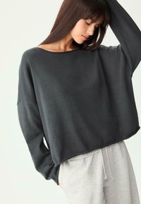Frau, die ein lockeres, dunkelgraues Langarmoberteil und eine hellgraue Jogginghose mit Kordelzug trägt, mit einem erhobenen Arm und nach unten blickend.