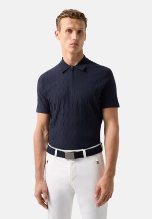 Mand iført tekstureret marineblå kortærmet polo med lynlås og hvide bukser, med et navyblåt og hvidt bælte med firkantet spænde.