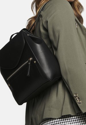 Donna che indossa un blazer verde oliva e pantaloni a fantasia pied de poule, con uno zaino in pelle nera con tasca anteriore con cerniera e patta.