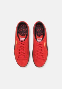 Crvene suédne tenisice s crnim detaljima, s teksturiranom potplatom, dizajnom na vezanje i uzorkom unutar. PUMA logo na jeziku i strani.