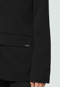 Schwarze Blazer mit strukturiertem Design, ausgestattet mit einer glatten Textur, umgeschlagenem Kragen und einem dezenten Akzent auf der Tasche mit einem metallischen Logo.