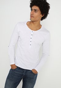 Langärmliges weißes Baumwollhemd mit rundem Ausschnitt und Knopfleiste. Mit grauen Knöpfen und taillierten Ärmeln. Kombiniert mit blauen Jeans.