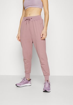 Femme portant un pantalon de jogging côtelé mauve, un soutien-gorge de sport mauve assorti et des chaussures de sport mauves, debout devant un fond blanc uni.