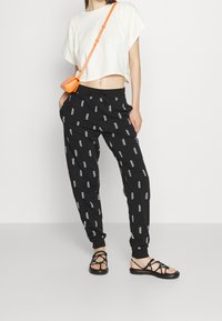 Zwarte joggerbroek met witte "HUGO" logo-print, cropped witte t-shirt en zwarte sandalen met bandjes. Model poseert met een oranje crossbody tas.