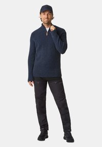Marineblå strikket pullover med halvlukning og træzipper; båret med sorte bukser med lynlås-lommer og en sort kasket.