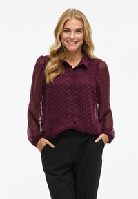 Blouse bourgogne à boutons sur le devant, tissu texturé et manches longues en voile ; présente un motif à pois discret et une coupe décontractée.