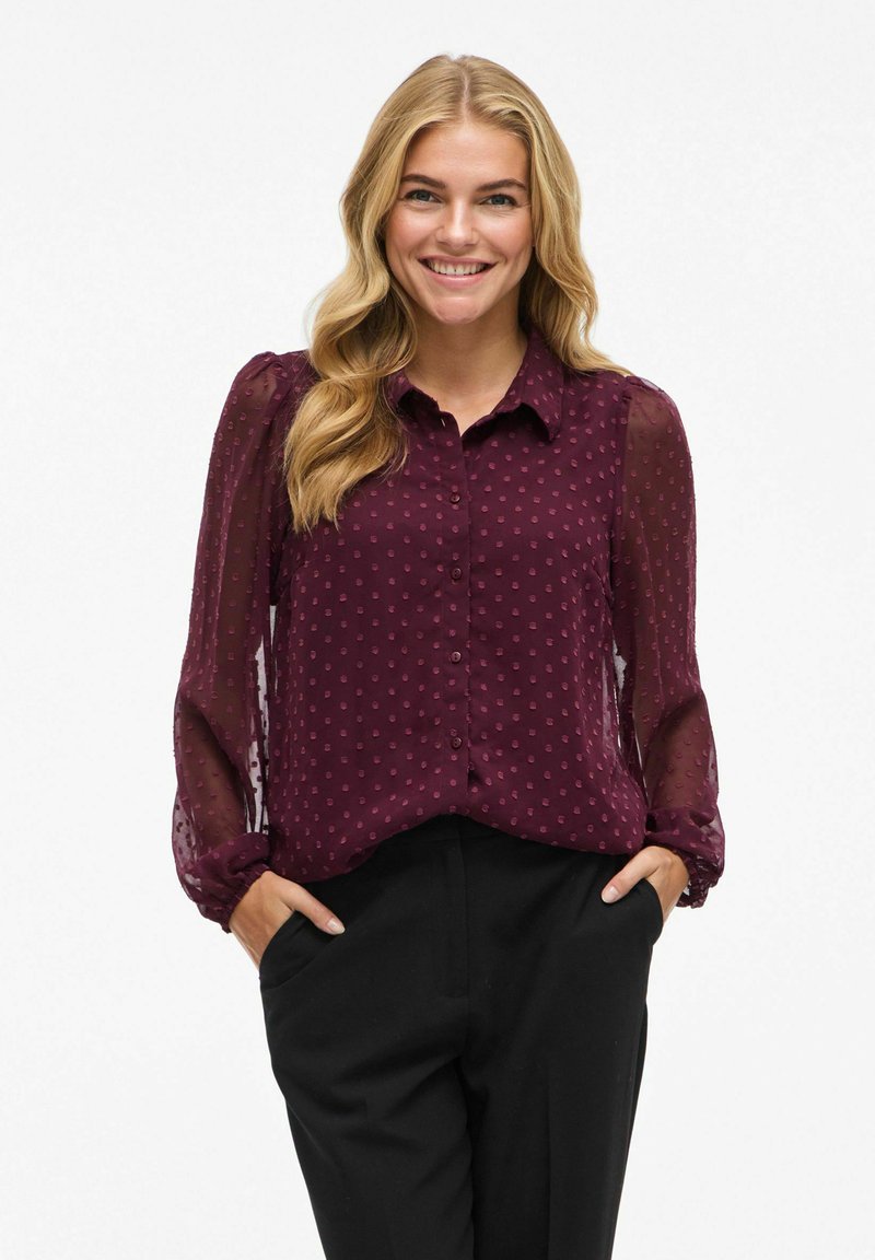 Blouse bourgogne à boutons sur le devant, tissu texturé et manches longues en voile ; présente un motif à pois discret et une coupe décontractée.