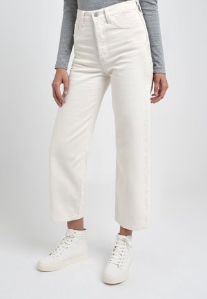 Personne portant un jean large taille haute couleur écru avec bouton et fermeture éclair à l'avant, associé à des baskets montantes blanches et un haut gris à manches longues.
