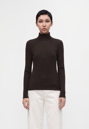 Person mit kurzen dunklen Haaren, die einen schwarzen Rollkragenpullover und weiße Hosen trägt, steht vor einem schlichten hellen Hintergrund.
