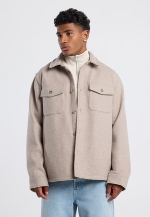Beige oversized wollen jas met twee borstzakken en knoopsluitingen, gedragen over een crème hoge hals top, gecombineerd met lichtblauwe jeans.