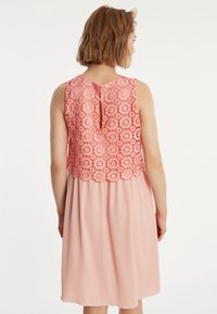 Korallenrosa Kleid mit einem Spitzenoberteil mit floralem Muster, einem Schlüsselloch-Ausschnitt im Rücken und einem fließenden Rock aus einem helleren rosa Stoff.