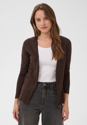 Jeune femme aux longs cheveux bruns portant un cardigan marron ouvert, un haut blanc et un jean taille haute gris foncé, debout devant un fond uni.