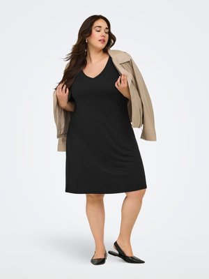 Kvinde iført en sort knælængde kjole og beige jakke hængende over skuldrene, med sorte slingback-sko på en hvid baggrund.