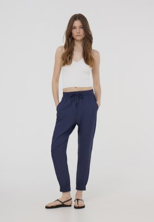 Pantaloni jogger blu navy con polsini elasticizzati, vita con cordoncino e tessuto leggero. Abbinati a una canotta bianca a coste.