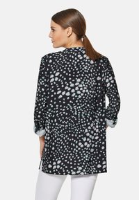 Schwarze Tunika mit einem bedruckten Muster aus helltürkisen und weißen Punkten. Lange Ärmel mit hochrollbaren Manschetten, lockere Passform und weicher Stoff.