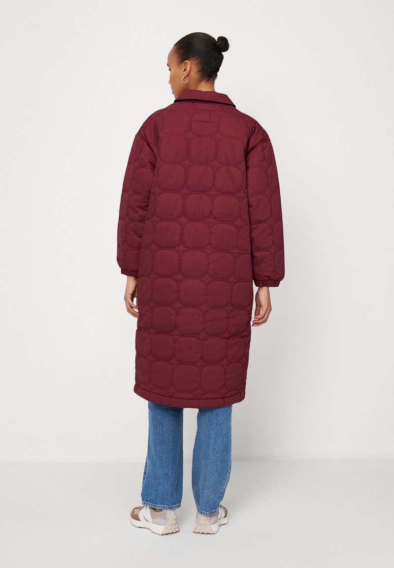 Manteau long matelassé bordeaux avec un motif de quilting circulaire, col à revers et épaules tombantes, associé à un jean bleu clair et des baskets blanches.