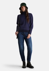 Pull en tricot bleu marine, côtelé au bas et aux poignets. Assorti avec un jean bleu et des bottes noires à lacets. Chapeau seau vert et petit sac bandoulière noir.