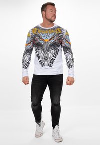 Langärmliges Grafik-T-Shirt mit einem auffälligen Adlerdesign in lebendigen Farben auf weißem Grund, mit schwarzer Schrift und taillierter Silhouette.