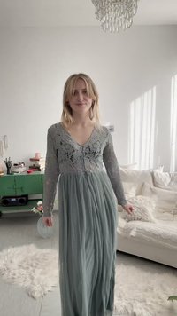 Junge Frau lächelnd, trägt ein langes graues Spitzenkleid, steht in einem sonnendurchfluteten Wohnzimmer mit weißem Sofa und grünem Schrank.