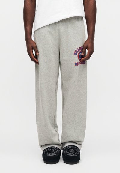 Γκρι βαμβακερή φόρμα με φαρδιά εφαρμογή, με κόκκινο και μπλε γραφικό σχέδιο στο αριστερό μηρό. Φοράει με μαύρες κεντημένες παντόφλες.