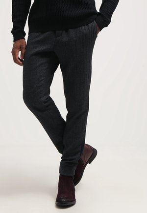 Mężczyzna w ciemnoszarych teksturowanych spodniach, czarnym swetrze i bordowych skórzanych botkach, stojący z jedną ręką w kieszeni i jedną nogą skrzyżowaną.