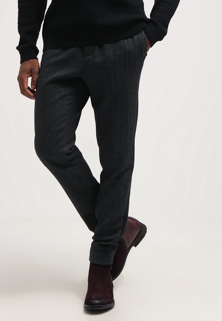 Homme portant un pantalon gris foncé texturé, un pull noir et des bottes en cuir bordeaux, debout, une main dans la poche et une jambe croisée.