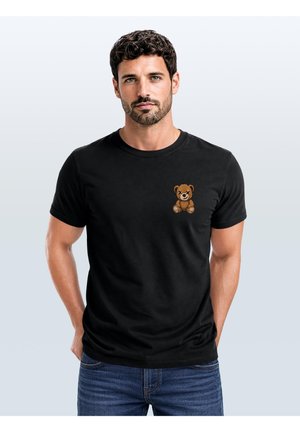 Man with short dark hair wearing black t-shirt with small teddy bear graphic on chest and blue jeans, standing with hands in pockets.