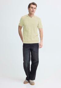 Man staat tegen een effen achtergrond, draagt een kort mouws, lichtgeel gebreid poloshirt, donkergelige losse spijkerbroek en lichtbruine sneakers met rubberen zolen.