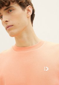 Brzoskwiniowy sweter z miękkiej bawełny, z okrągłym dekoltem i małym białym logo na lewej piersi.