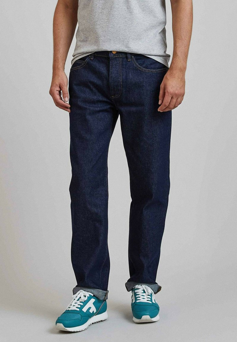 Donkerblauwe spijkerbroek met een rechte pijp, gecombineerd met een grijs T-shirt en teal sneakers met witte zolen en veters.